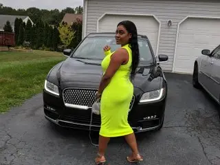 BrownRound21 poses next                    tto a black car wearing a tight neon yellow tank top and shorts, letting her long dark hair hang down her back.