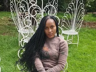 Browncsndy018 poses outdoors with long black hair, wearing a brown long-sleeved top while sitting in a white decorative metal chair.