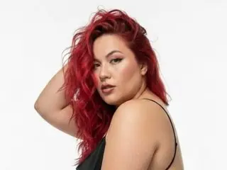 BrujaSCarlata poses with her long red hair, showing her bare shoulders and chest in a close-up shot against a white background.