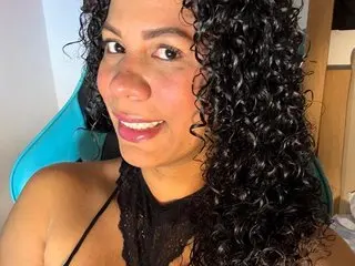 Smiling woman with long curly black hair wearing a black lace top in a bright indoor setting.