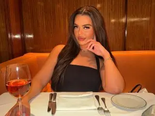 Brunette_bunnyxo poses with long dark hair, wearing a black tank top while sitting at a table with a glass of red wine.