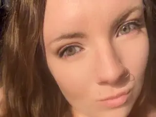 BustBella31 looks directly at the camera with her long brown hair framing her face in a close-up shot.