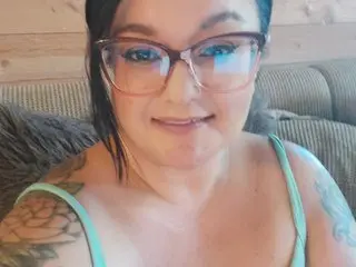BustyFarmBabe looks at the camera with a smile, wearing glasses and a light mint green camisole, showing her shoulders and collarbone.
