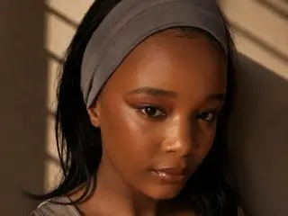 BustyM_XX poses with a direct gaze, showing her face and dark hair against a warm, sunlit background.