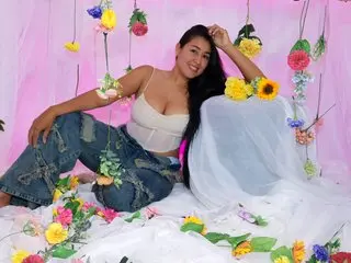 Busty_May poses with long dark hair, wearing a white tank top and denim jeans while sitting on a white bedsheet surrounded by colorful flowers.