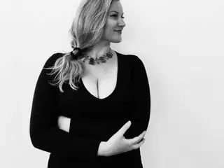 Bustybeauty18 poses against a white background, wearing a black long-sleeved top that shows deep cleavage, with her blonde hair styled down.