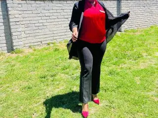Bustypillows20 stands outside wearing a red top and black pants, posing with her hands at her sides against a brick wall and green grass.