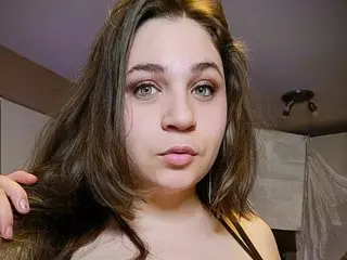 Bustyprincessleila looks at the camera with long brown hair and a direct gaze, wearing a black top against a plain indoor background.