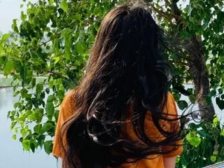 Bustysweta poses outdoors with her long, dark hair flowing down her back, wearing an orange top against a background of green leaves.