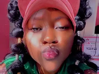 ButtyshanXxx wearing a red cap and green shirt, making a kissing face with her dark braided hair.