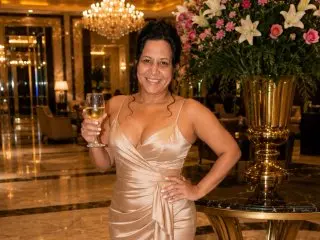 Camil4milf smiles while wearing a low-cut, silky gold slip dress, posing in a luxury hotel setting with a glass of white wine.