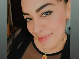 Smiling woman with winged eyeliner and gold necklace wearing a black top in a soft indoor setting.