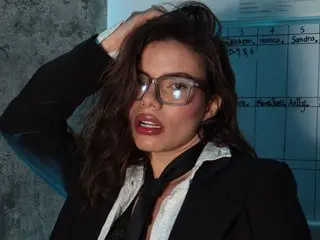 Confident brunette woman in professional black blazer and glasses posing in a modern office setting.