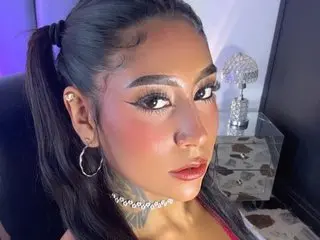 CandyMarilyn poses with her dark hair in a ponytail, wearing a silver choker and heavy eye makeup against a plain background.