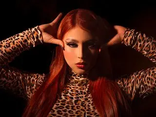 CanndyCoox poses with her hands behind her head, wearing a long-sleeved leopard print top that shows her long red hair and face.