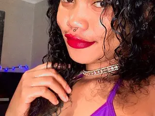 Cardibabe21 shows off her long curly hair and red lips while wearing a silver choker and a purple top.