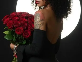CarlaMercer poses in a black dress while holding a large bouquet of red roses against a bright circular light.