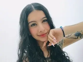 Carla_Cortes smiles while posing with her hand near her face, showing her long, dark wavy hair against a plain white background.