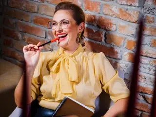 CaroJhones smiles wearing a yellow tie-neck blouse and glasses, posing against a rustic brick wall background.