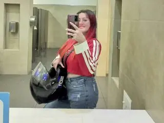 Woman in red crop top and blue jeans taking a mirror selfie in a restroom while holding a black motorcycle helmet.