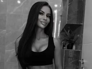 CarollineFoxy poses with long dark hair, wearing a black tank top against a light-colored wall background.
