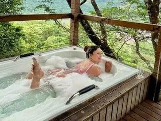 Relaxed woman in a pink bikini enjoying a bubbly outdoor hot tub overlooking a lush green forest landscape.