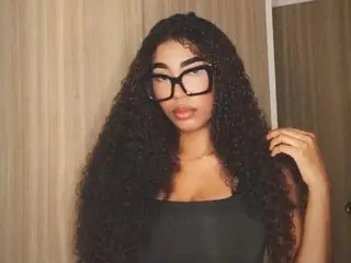 CataFooxy30 poses with her long, dark curly hair down, wearing black glasses and a black tank top against a neutral wall.