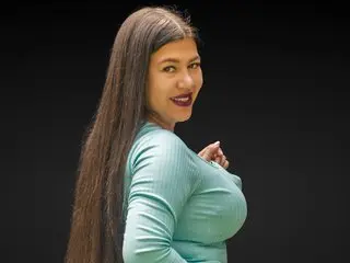 Smiling woman with long dark hair wearing a light blue ribbed top against a black background.