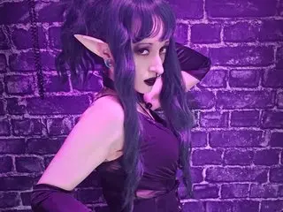 Cathulhu, with long purple hair and a black top, poses against a purple brick wall.
