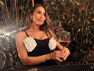 CatifeaRosie poses with long brown hair, wearing a black tank top with white ruffles, holding a glass of wine in a dimly lit setting.