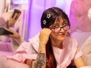 Celestewang poses with her hands near her face, wearing glasses and a light pink top against a soft purple background.