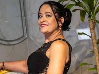 Smiling woman in a black sequined evening dress posing indoors near a green houseplant.