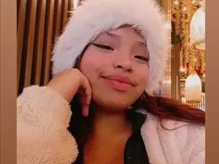 CharlotAura poses with her hand under her chin, wearing a white fluffy hat and a white fuzzy jacket against a warm, lit background.