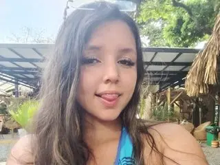 CharlotteWhitmore with long brown hair, looking at the camera while sticking her tongue out slightly in an outdoor setting.