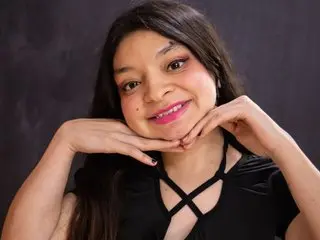 Smiling young woman with long dark hair wearing a black crisscross top against a dark studio background.