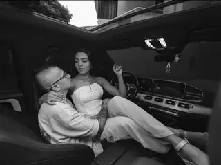 CharlotteandSimon, a woman with dark hair, poses in a car wearing a white bustier and white pants, looking towards the camera.