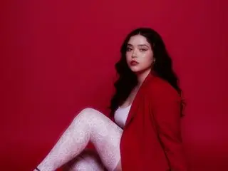 Charlottesinclair22 poses with long dark hair, wearing a red blazer and white lace leggings against a red background.