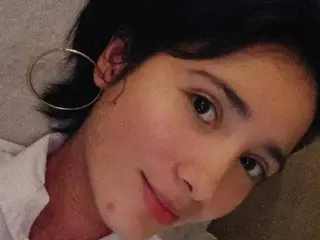 Cherrylb with short dark hair and large hoop earrings smiles while laying down in a white shirt.