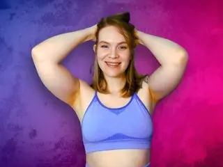 ChillBillie smiles at the camera while posing with her hands in her hair, wearing a blue sports bra against a purple and pink background.