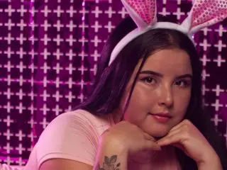 ChloeFlorez wearing pink bunny ears and a pink top while posing against a purple background.