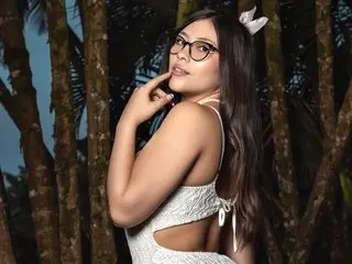 ChloeFrey poses with her hand near her face, showing her long dark hair and white lace outfit against a dark tropical background.