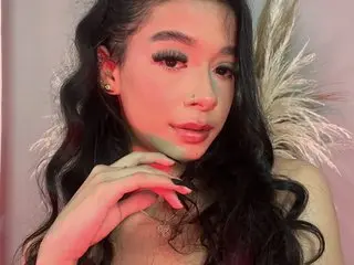 ChloeRosses poses with long wavy dark hair, showing her face and shoulders against a soft pink background.