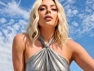 ChloeVee poses in a silver halter top against a clear blue sky, showing her blonde hair and bare shoulders.