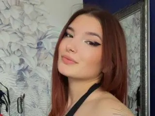ChloeXSmith poses with a soft smile, showing her long reddish-brown hair and clear skin against a light floral background.