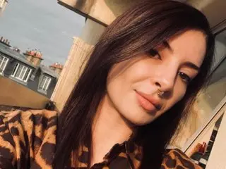 ChloeXsecret poses with long dark hair, wearing a brown patterned top against a bright outdoor background.