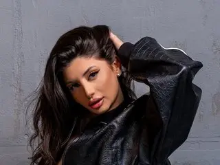 Chloe_Sin poses with her hands in her long dark hair, wearing a black leather top against a grey wall.