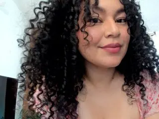 ChubbyLicius poses with her long, dark curly hair, wearing a pink floral top against a white wall.