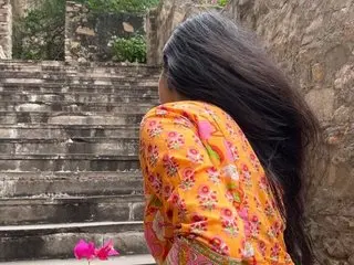 ChubbyShweta poses with her long black hair flowing down her back while wearing a bright orange floral patterned top outdoors near stone steps.