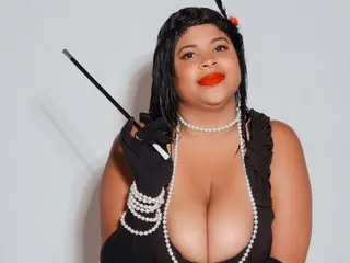 Glamorous woman in a black evening dress and pearls holding a vintage whip against a plain background.
