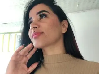 CinthiaCM looks at the camera with long dark hair and a tan turtleneck, resting her hand against her neck.